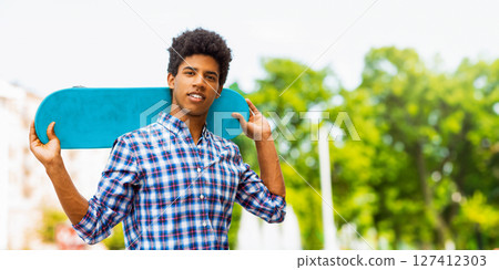 A teen black guy with dark curly hair and a plaid shirt is smiling while carrying a blue skateboard on his shoulder. The background shows a blurred view of trees and buildings, copy space A teen black guy with dark curly hair and a plaid shirt is smiling while carrying a blue skateboard on his shoulder. The background shows a blurred view of trees and buildings, copy space 127412303