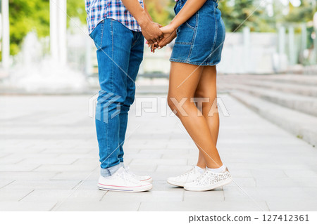 Unrecognizable couple holding hands on a city street. They are both wearing jeans, white sneakers, and casual clothing. Guy is wearing a plaid shirt and the girl is wearing a denim skirt, cropped 127412361