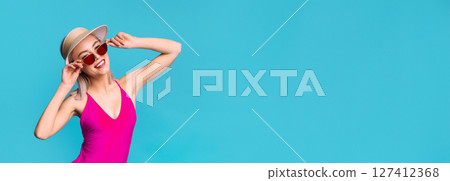 A young woman wearing a pink swimsuit, a straw hat, and sunglasses smiles and poses while standing against a blue background. The woman appears to be in a carefree and happy mood, copy space A young woman wearing a pink swimsuit, a straw hat, and sunglasses smiles and poses while standing against a blue background. The woman appears to be in a carefree and happy mood, copy space 127412368
