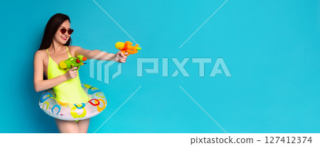 A woman in a bright yellow swimsuit and sunglasses holds two water guns, one in each hand. She has a playful expression on her face and is looking towards the right side of the frame A woman in a bright yellow swimsuit and sunglasses holds two water guns, one in each hand. She has a playful expression on her face and is looking towards the right side of the frame 127412374