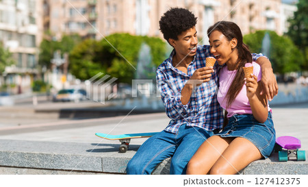 A teen black couple is sitting on a ledge in the city, enjoying ice cream cones on a sunny day. They are laughing and smiling, clearly enjoying each others company, copy space 127412375