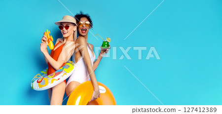 This image features two women in swimsuits posing against a blue background. Both women are smiling and look ready for a fun day at the pool, copy space 127412389