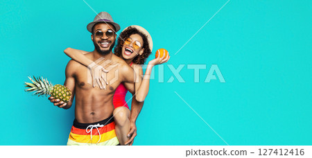 African American young couple is seen smiling and having fun in front of a bright turquoise background. The man wearing swim trunks and a hat, while the woman is in a swimsuit and holding an orange. 127412416
