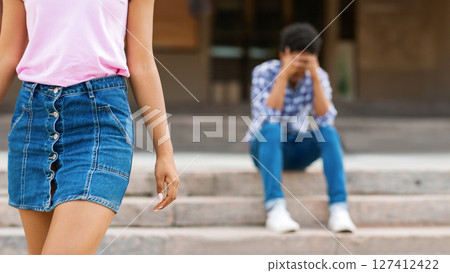 A girl in a pink shirt and denim skirt walks away from a guy sitting on concrete steps. Guy is wearing jeans and a plaid shirt. The girl is looking straight ahead while guy is looking down A girl in a pink shirt and denim skirt walks away from a guy sitting on concrete steps. Guy is wearing jeans and a plaid shirt. The girl is looking straight ahead while guy is looking down 127412422