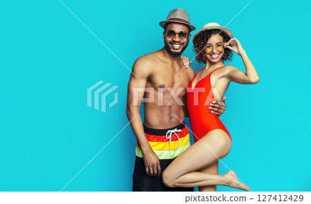 African American young couple wearing swimwear and hats, posing against a bright blue background. They both appear happy and relaxed, enjoying the summer weather. 127412429