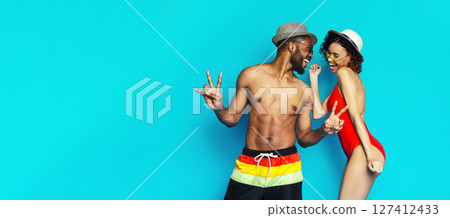 A young black couple in summer clothing are posing against a bright blue background. They are both smiling and appear to be enjoying each others company, copy space 127412433