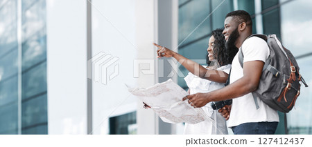 African American couple stand outside a building with modern architecture. The woman is pointing to the right, while the man is holding a map and smiling, copy space 127412437