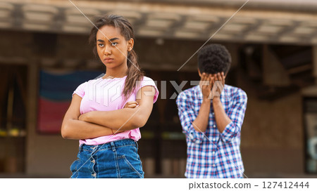 A teen black teenage girl stands outside a school building with her arms crossed and a stern expression. In the background, a teenage boy standing with his face in his hands, crying. 127412444