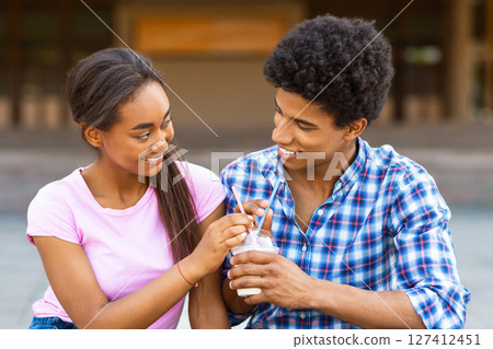 A teen black couple is sitting outside on a sunny day, sharing a drink with straws. They are both smiling and looking at each other. The couple appears to be enjoying their time together. 127412451