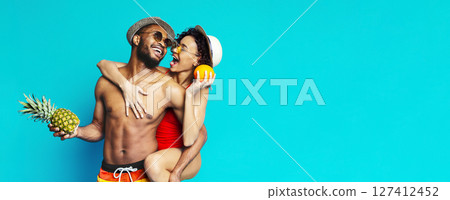 A young couple in swimwear poses in front of a blue background. The man is holding a pineapple and the woman is holding an orange. They are both smiling and looking at each other. 127412452