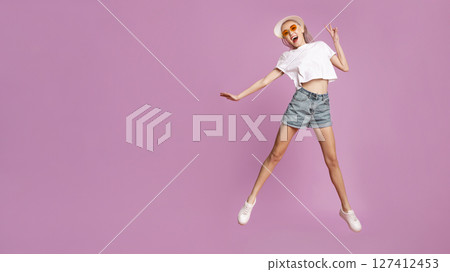 A young woman is captured mid-air, jumping with excitement against a pink backdrop. She is wearing a white t-shirt, denim shorts, and sunglasses, and is smiling broadly with her arms outstretched. 127412453