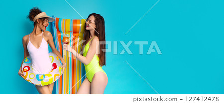 This image depicts two young women, one in a white swimsuit and the other in a green swimsuit, standing together in front of a bright blue background, copy space 127412478