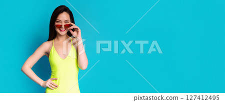 A young woman, with dark hair and a bright smile, is wearing a neon yellow swimsuit and red sunglasses. She is posing against a bright blue background, copy space 127412495