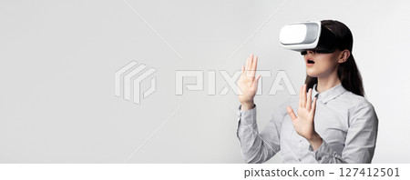 A woman wearing a virtual reality headset looks surprised as she reaches out her hands to touch something in the virtual world. A woman wearing a virtual reality headset looks surprised as she reaches out her hands to touch something in the virtual world. 127412501