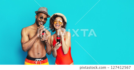 A young black couple dressed in swimwear and sunglasses smiles and enjoys drinks together on a bright blue background. 127412567