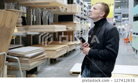 Contractor using a smartphone to select wood materials in a lumber warehouse for an upcoming project. A man in a hardware store, choosing lumber, shelves with lumber. Contractor using a smartphone to select wood materials in a lumber warehouse for an upcoming project. A man in a hardware store, choosing lumber, shelves with lumber. 127412638