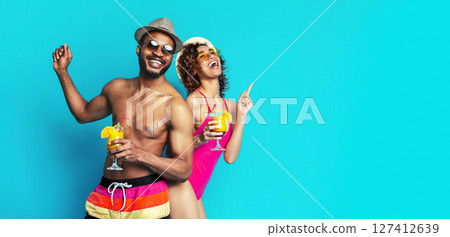 This image shows a man and a woman dressed in summer attire and smiling. They are both holding cocktails and appear to be enjoying themselves. The background is a vibrant blue. 127412639