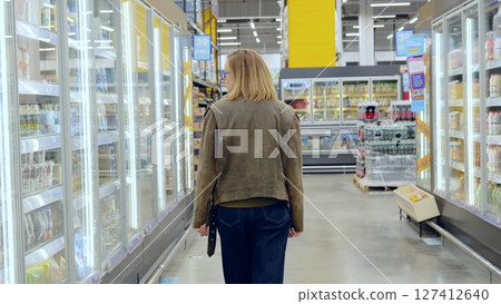Young woman strolling through the grocery store, carefully selecting items from the chilled display case Young woman strolling through the grocery store, carefully selecting items from the chilled display case 127412640