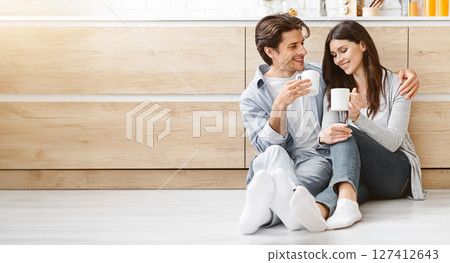 A young couple sits on the floor in a modern kitchen, enjoying a coffee break together. They are both smiling and looking at each other, enjoying their shared moment, copy space A young couple sits on the floor in a modern kitchen, enjoying a coffee break together. They are both smiling and looking at each other, enjoying their shared moment, copy space 127412643