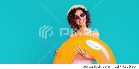 This image shows African American woman with curly hair wearing sunglasses and a straw hat, smiling and holding a bright orange inflatable ring against a turquoise background. 127412650
