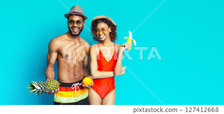 Black smiling couple in swimwear holding tropical fruit against a blue background. They are both wearing sunglasses and hats, and seem to be enjoying their summer day, copy space 127412668