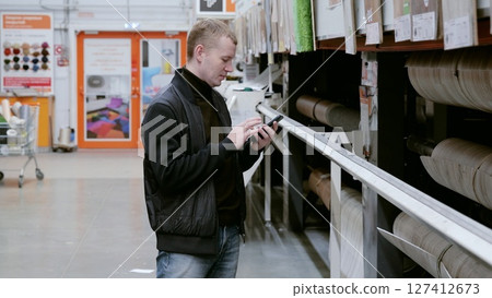 A man in a hardware store choosing flooring. Customer using a smartphone to compare prices, read reviews, and find more details about linoleum flooring while browsing in a hardware store 127412673