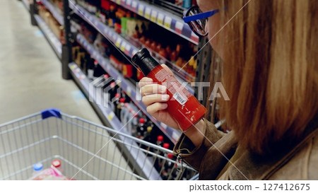 Customer carefully selects hot sauce while reading label and ingredients in a supermarket, holding a bottle of red hot sauce and pushing a shopping cart Customer carefully selects hot sauce while reading label and ingredients in a supermarket, holding a bottle of red hot sauce and pushing a shopping cart 127412675