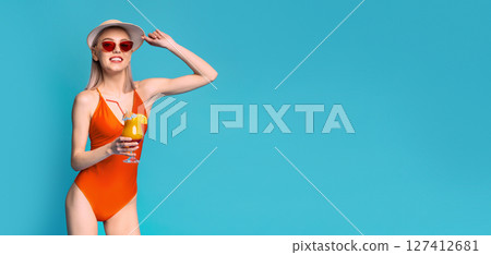 Young woman in an orange swimsuit, wearing sunglasses and a straw hat, holding a cocktail with a straw, standing in front of a solid blue background. She is looking at copy space 127412681