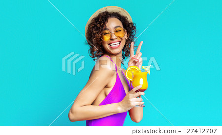 A young black woman with curly brown hair is wearing a purple swimsuit, a straw hat, and yellow sunglasses. She is smiling and holding up a peace sign while holding a tropical cocktail. 127412707