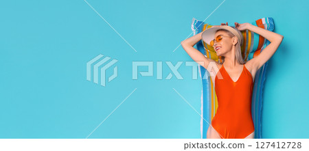A young woman wearing a red swimsuit, sunglasses, and a straw hat is lying on a colorful inflatable raft, smiling and relaxing on a blue background. She has a carefree and summery expression. 127412728