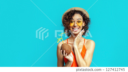 African American woman in a swimsuit, straw hat, and sunglasses is holding a coconut drink with a straw. She is smiling brightly and looking directly at the camera. The background is blue African American woman in a swimsuit, straw hat, and sunglasses is holding a coconut drink with a straw. She is smiling brightly and looking directly at the camera. The background is blue 127412745