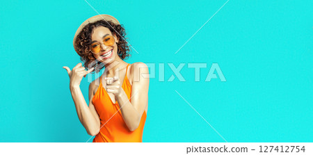 A young black woman with curly hair is wearing a bright orange swimsuit, a straw hat, and yellow sunglasses. She is posing against a bright turquoise background and smiling broadly, copy space A young black woman with curly hair is wearing a bright orange swimsuit, a straw hat, and yellow sunglasses. She is posing against a bright turquoise background and smiling broadly, copy space 127412754