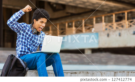 A teen black guy sits on a concrete ledge outside a building, celebrating a victory on his laptop. He has a wide smile on his face and his fist raised in the air, showing his excitement, copy space A teen black guy sits on a concrete ledge outside a building, celebrating a victory on his laptop. He has a wide smile on his face and his fist raised in the air, showing his excitement, copy space 127412774