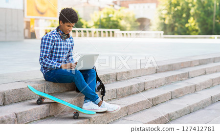 This image shows a teen black guy sitting on a set of stone steps in a city environment. He is using a laptop and has a skateboard beside him, copy space This image shows a teen black guy sitting on a set of stone steps in a city environment. He is using a laptop and has a skateboard beside him, copy space 127412781