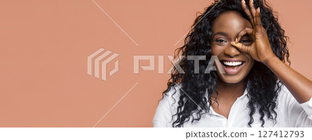 Black happy woman with long, curly black hair. She is wearing a white shirt and smiling broadly while gesturing with her hand in front of her eye, creating an okay symbol 127412793