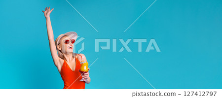 Happy woman in a red swimsuit and a white sunhat, with her arm raised in the air, holding a cocktail in her other hand. She is looking to the side with a big smile on her face, copy space 127412797