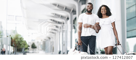 Black couple walks hand-in-hand through an airport terminal, both smiling and looking happy. They are carrying luggage and appear to be excited for their trip, copy space 127412807