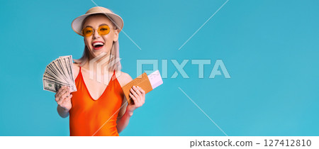 A young woman wearing a straw hat, orange swimsuit, and sunglasses smiles and holds a fan of cash and a boarding pass. She is standing in front of a bright blue background, copy space 127412810
