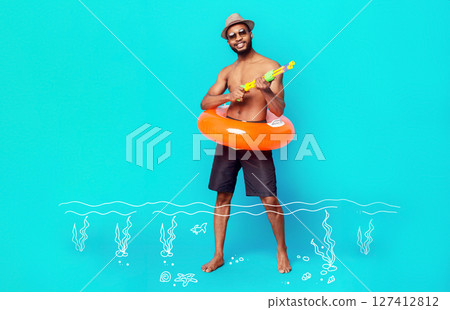 Joyful black guy with water gun and inflatable ring posing over blue studio background, enjoying his summer vacation 127412812