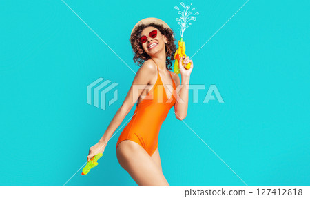 Black woman in a bright orange swimsuit smiles brightly while holding two water guns. She wears a straw hat and red sunglasses. The background is a vibrant blue Black woman in a bright orange swimsuit smiles brightly while holding two water guns. She wears a straw hat and red sunglasses. The background is a vibrant blue 127412818