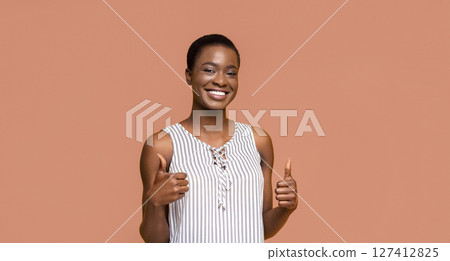 A young Black woman with short hair is smiling and giving a thumbs up with both hands. She is wearing a white and black striped sleeveless top and standing in front of a peach-colored background. A young Black woman with short hair is smiling and giving a thumbs up with both hands. She is wearing a white and black striped sleeveless top and standing in front of a peach-colored background. 127412825