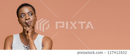 African American woman with short, dark hair stands against a peach-colored background. Her finger rests under her chin as she looks off to the left, appearing thoughtful. African American woman with short, dark hair stands against a peach-colored background. Her finger rests under her chin as she looks off to the left, appearing thoughtful. 127412832