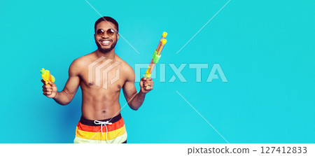 This image features black man wearing sunglasses and swim trunks, holding two water guns in his hands. He is smiling and looking towards the camera. The background is a solid blue color. This image features black man wearing sunglasses and swim trunks, holding two water guns in his hands. He is smiling and looking towards the camera. The background is a solid blue color. 127412833