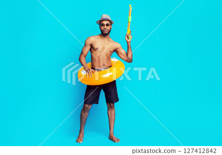 Black man wearing a hat, sunglasses, and swim trunks stands in front of a blue background. He is holding a yellow water gun in his right hand and an inflatable ring around his waist. 127412842