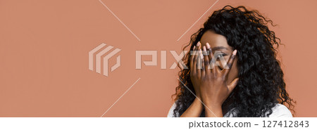 This is a photo of a young Black woman with long, curly hair standing against a tan background. She is smiling and covering her face with both hands. 127412843