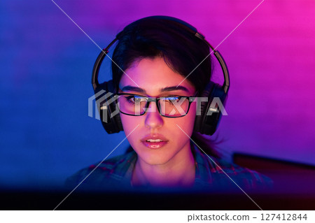 A young woman with dark hair, wearing headphones and glasses, sits in front of a computer screen. She appears to be focused on the screen, with a serious expression on her face 127412844