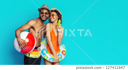 Black couple posing in front of a blue background. The man is holding a red and white beach ball, while the woman is holding a green water gun and a colorful inflatable ring, copy space 127412847