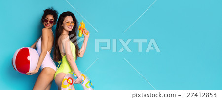 This image shows two young women posing in swimsuits against a bright blue background. One woman is holding a large beach ball, while the other is holding a water gun, copy space 127412853