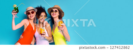 Three friends, wearing swimsuits, hats, and sunglasses, smile brightly as they hold cocktails in front of a blue background. The woman on the left holds a green cocktail with a straw, copy space Three friends, wearing swimsuits, hats, and sunglasses, smile brightly as they hold cocktails in front of a blue background. The woman on the left holds a green cocktail with a straw, copy space 127412862