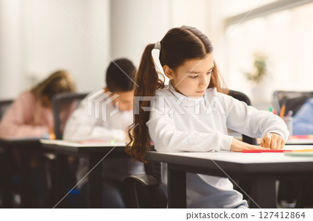 Portrait of small girl with ponytails sitting at table in classroom at primary school or kindergarten, writing or drawing in notebook. Reopening and return back to school after coronavirus quarantine 127412864